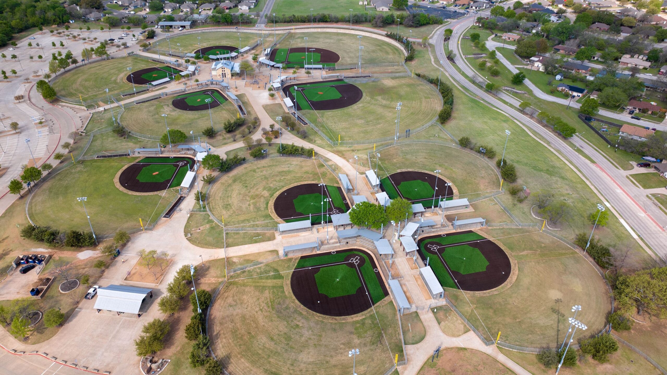 Aerial view of a sports complex featuring multiple circular baseball fields with green artificial turf and brown infields, surrounded by trees and roads.